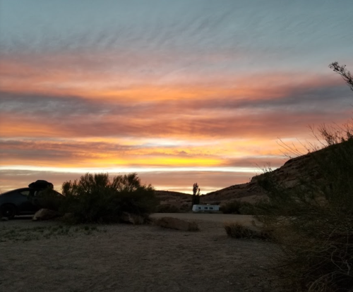 A vibrant sunset paints the sky with orange and purple hues over a desert landscape with sparse vegetation.